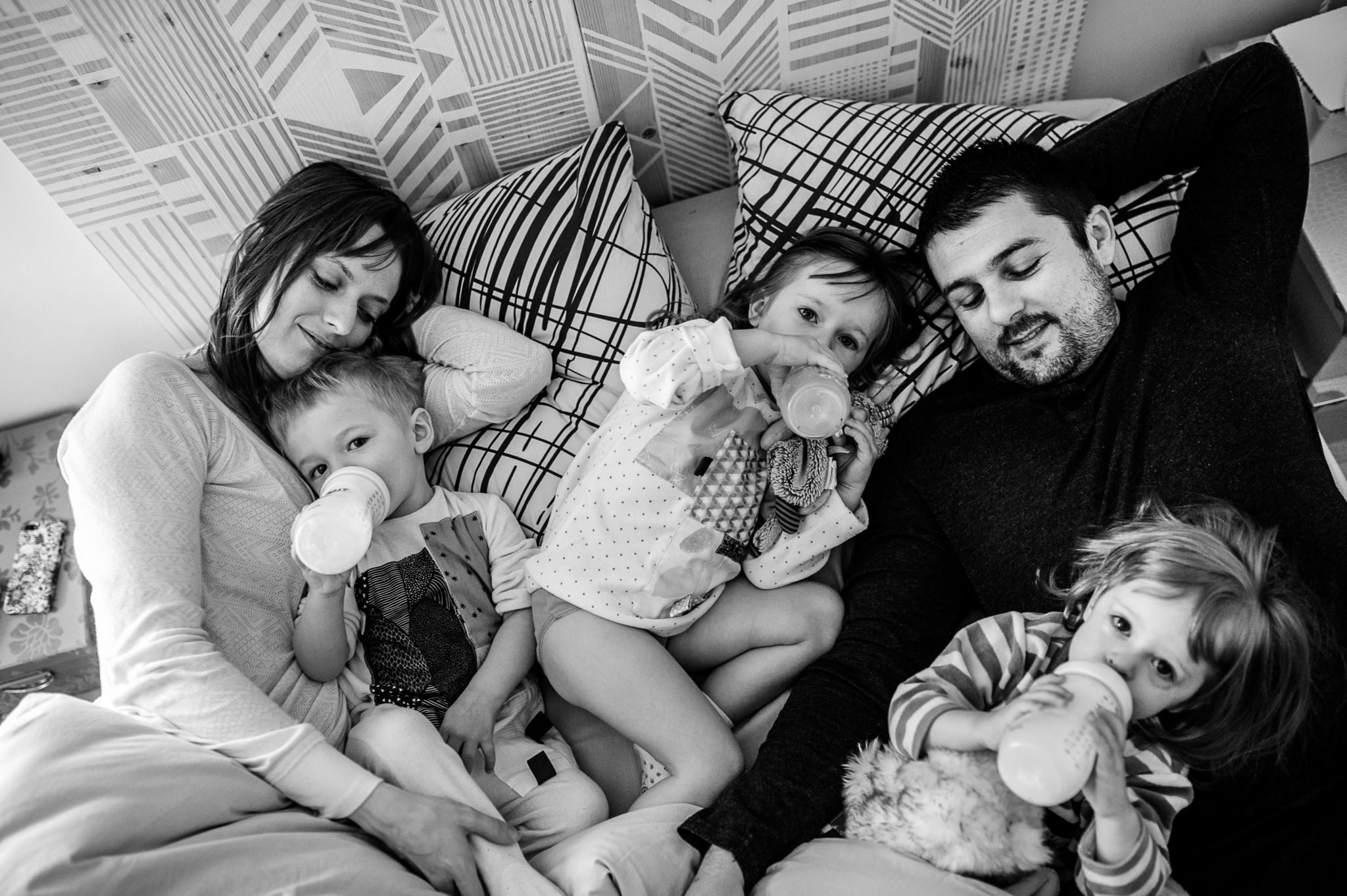Famille avec trois jeunes enfants buvant leur biberon du matin, allongés dans le lit des parents