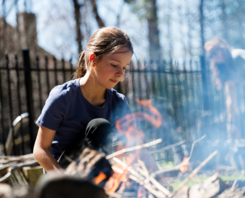 Jeune fille concentrée sur les flammes d'un feu de bois dans la cour, à travers la fumée