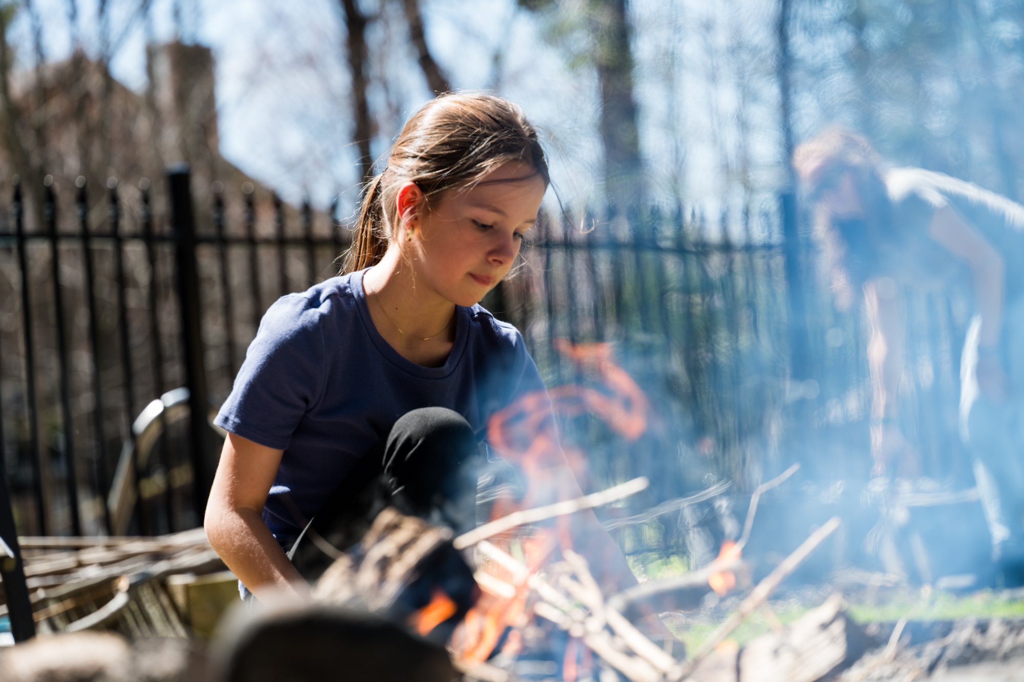 Jeune fille concentrée sur les flammes d'un feu de bois dans la cour, à travers la fumée
