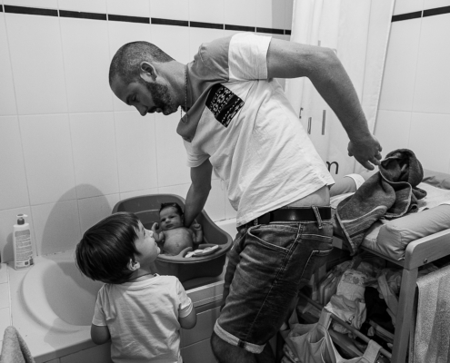 Père donnant le bain à son nouveau-né pendant que son aîné observe la scène, en noir et blanc
