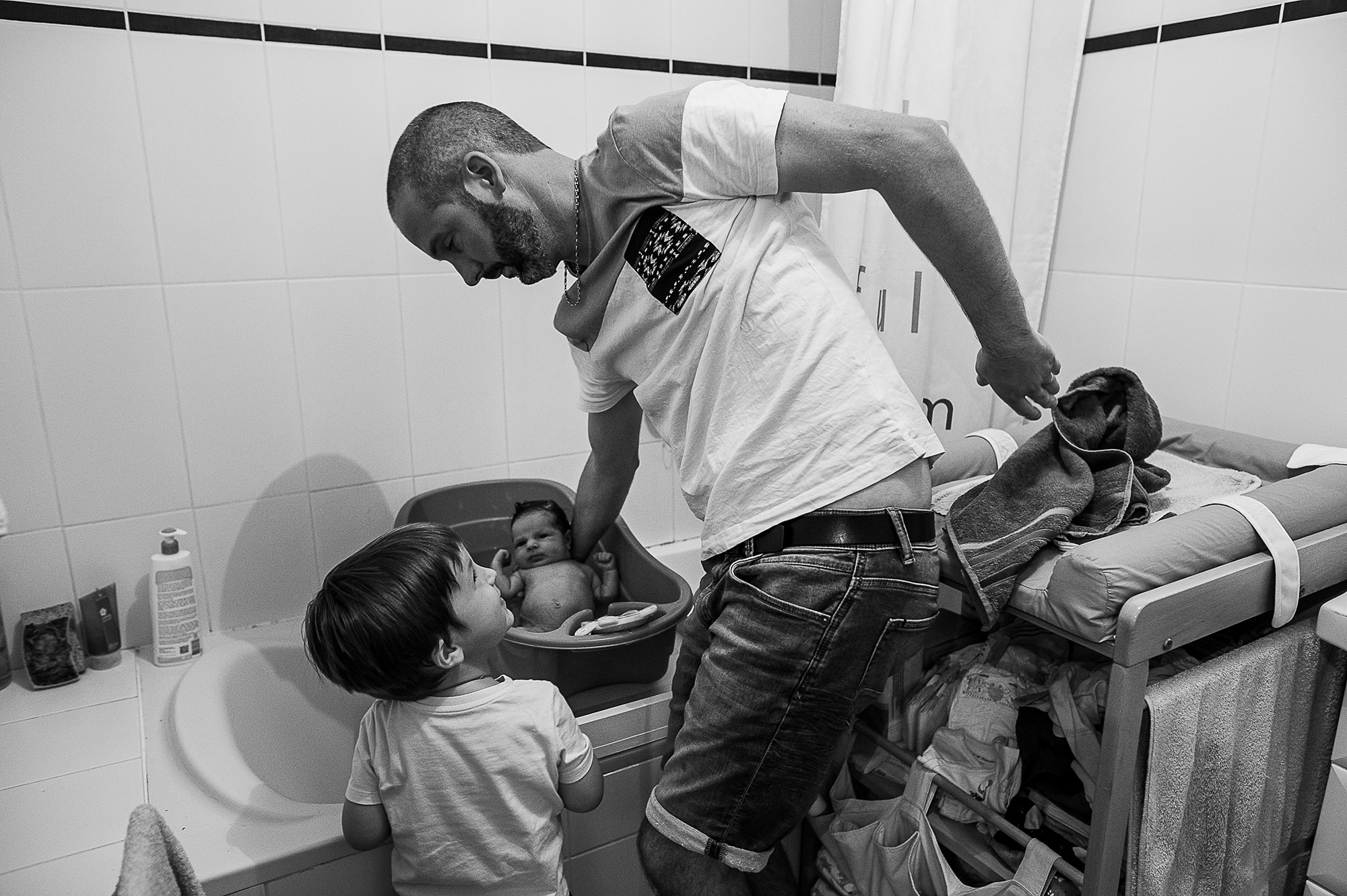 Père donnant le bain à son nouveau-né pendant que son aîné observe la scène, en noir et blanc