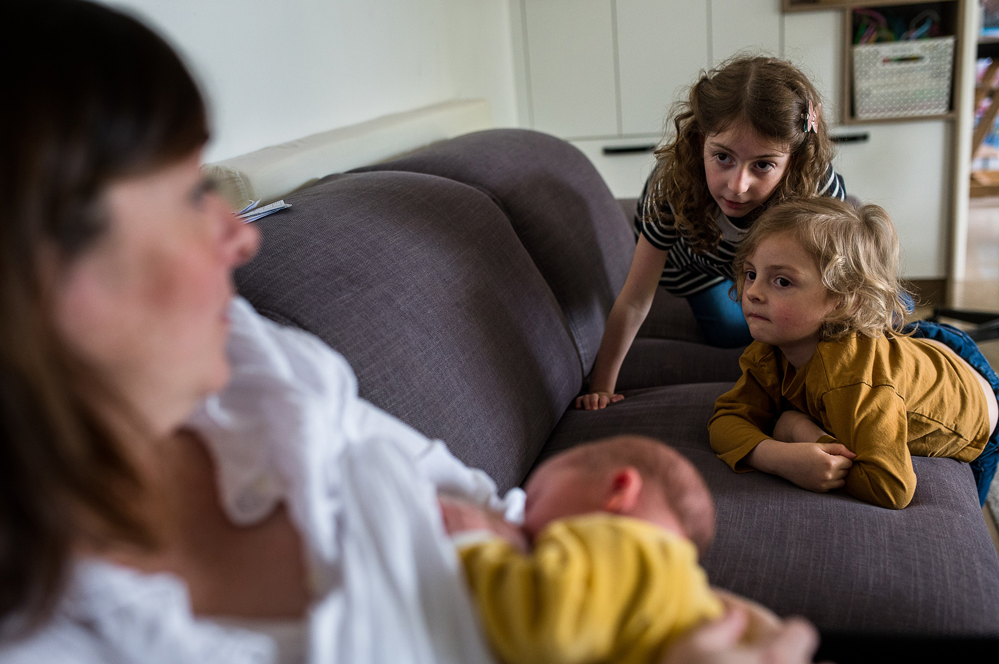 Deux aînées appuyées contre le dossier du canapé, observant attentivement leur nouveau-né petit frère ou petite sœur dans les bras de leur mère