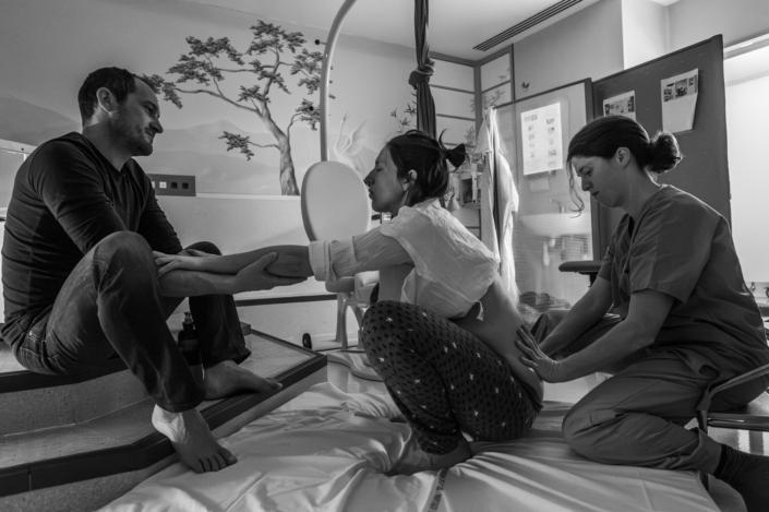 Femme en travail accroupie au sol dans une salle de naissance, elle tient la main de son conjoint assis en face d'elle, une sage-femme applique une contre-pression sur son bassin