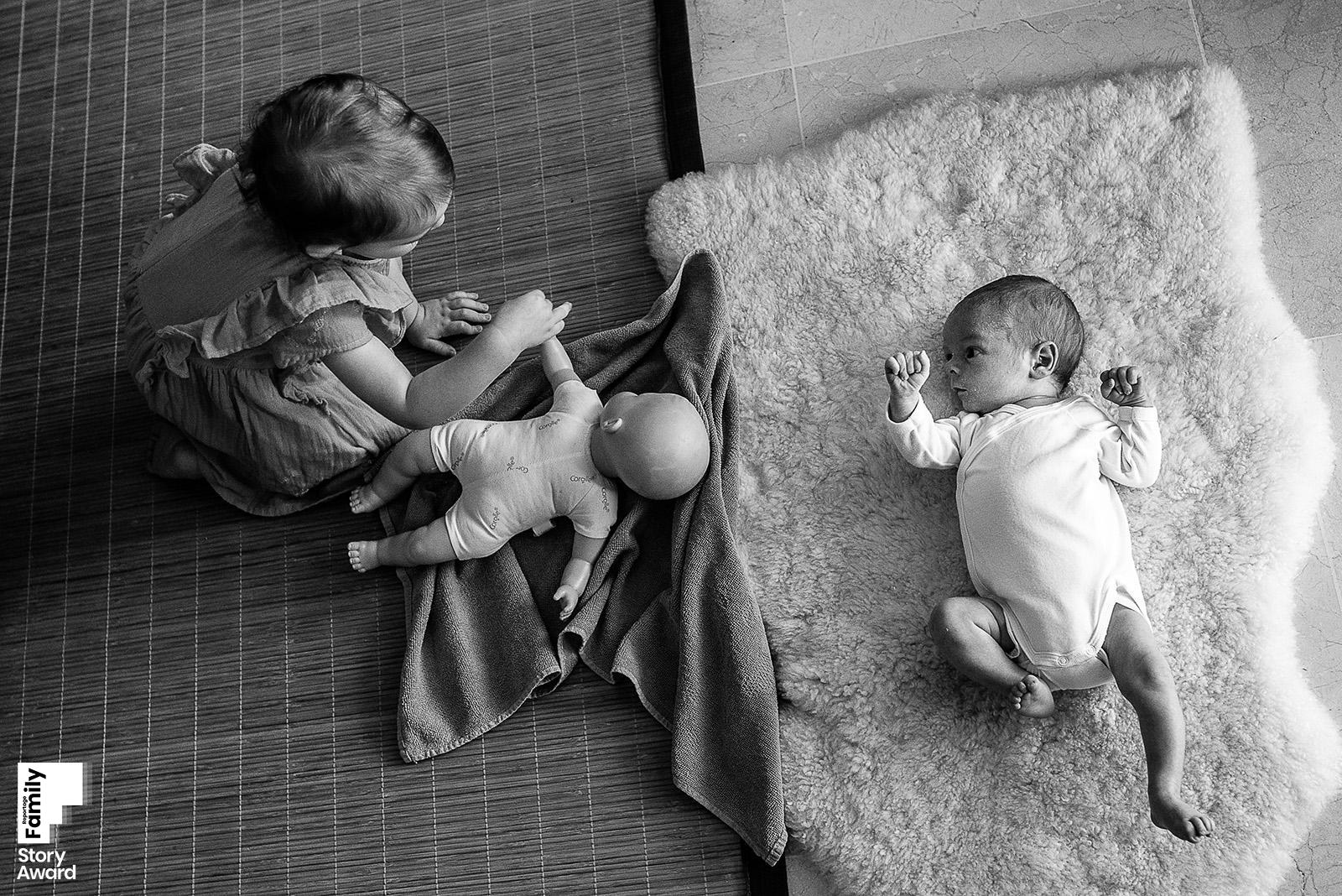 Petite fille assise sur un tatami joue à la poupée à côté de son petit frère nouveau-né allongé sur un tapis de peau de mouton, vue plongée en noir et blanc